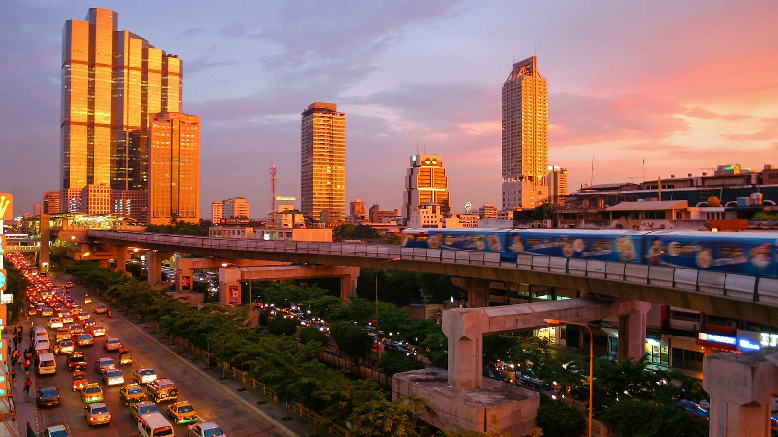 夕陽下的曼谷市區，BTS 空鐵高架橫過馬路，橋下是滿滿的車陣，遠方是市中心的高樓天際線