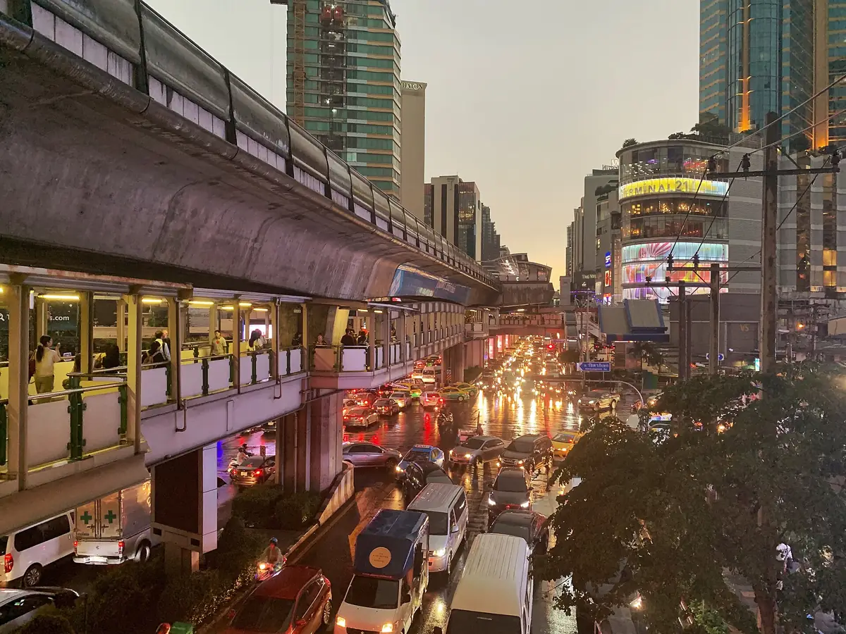 傍晚的蘇坤蔚路阿索克路口，BTS 空鐵高架沿街延伸，橋下是滿滿的車流與亮燈的商場
