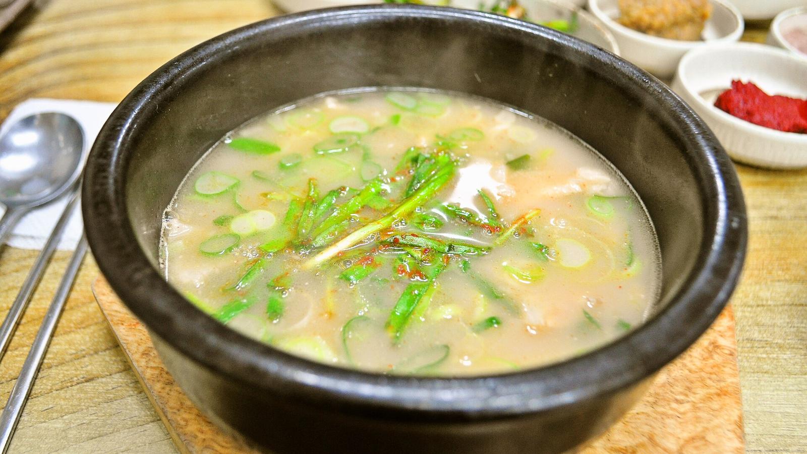黑色石鍋裝著豬肉湯飯，米白色的湯上鋪滿翠綠蔥花與韭菜，湯裡看得到薄切豬肉，後方桌上擺著幾碟小菜