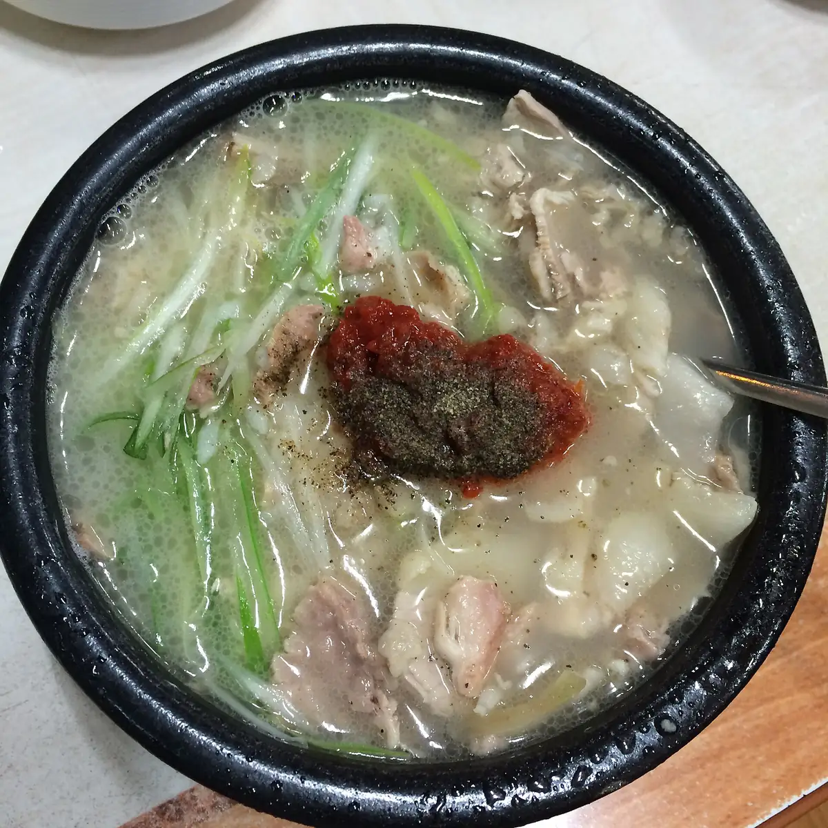 俯拍一碗豬肉湯飯，米白色的湯中央放了一大匙鮮紅色辣醬，上面撒著深色粉末，旁邊看得到切成細絲的青蔥與豬肉片，一支湯匙從右側伸入碗中