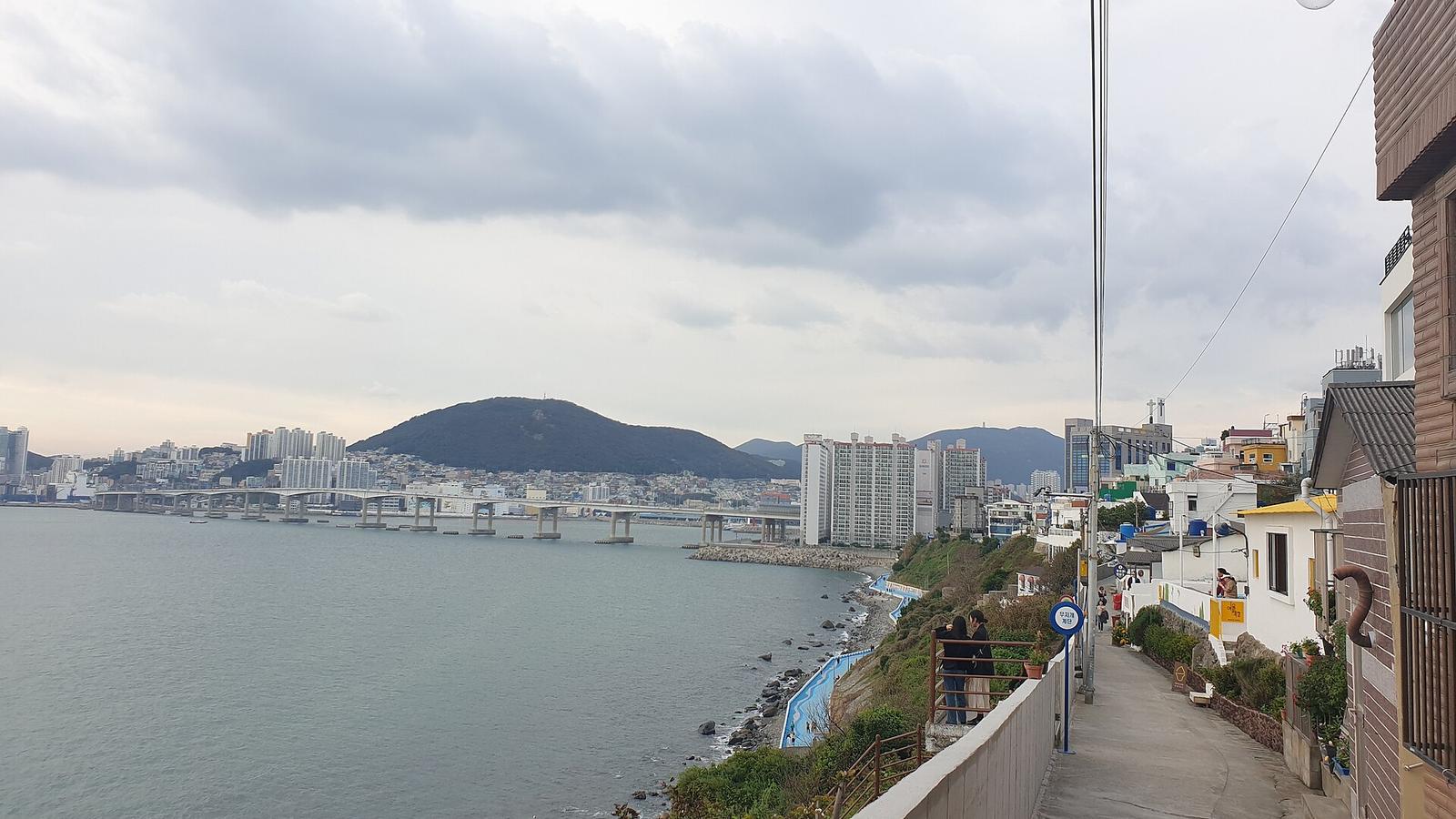 沿著海岸山壁而建的白色矮房聚落，窄巷步道面向海灣，遠方有跨海大橋與城市天際線