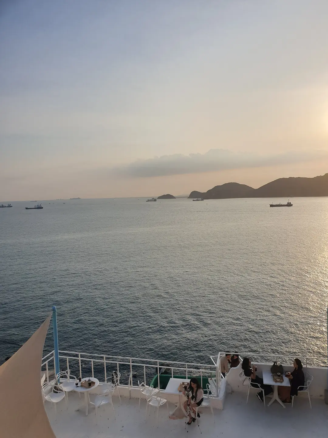 夕陽下的海景露臺咖啡座,白色桌椅面向大海,遠方海面上有幾座島嶼與貨輪剪影