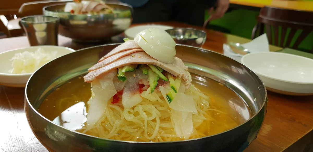 桌上一碗小麥冷麵，麵上鋪著切片肉、小黃瓜絲、白蘿蔔片與一塊切開的水煮蛋，湯是淺褐色，背景可見另一碗同樣的麵與幾個小菜碟