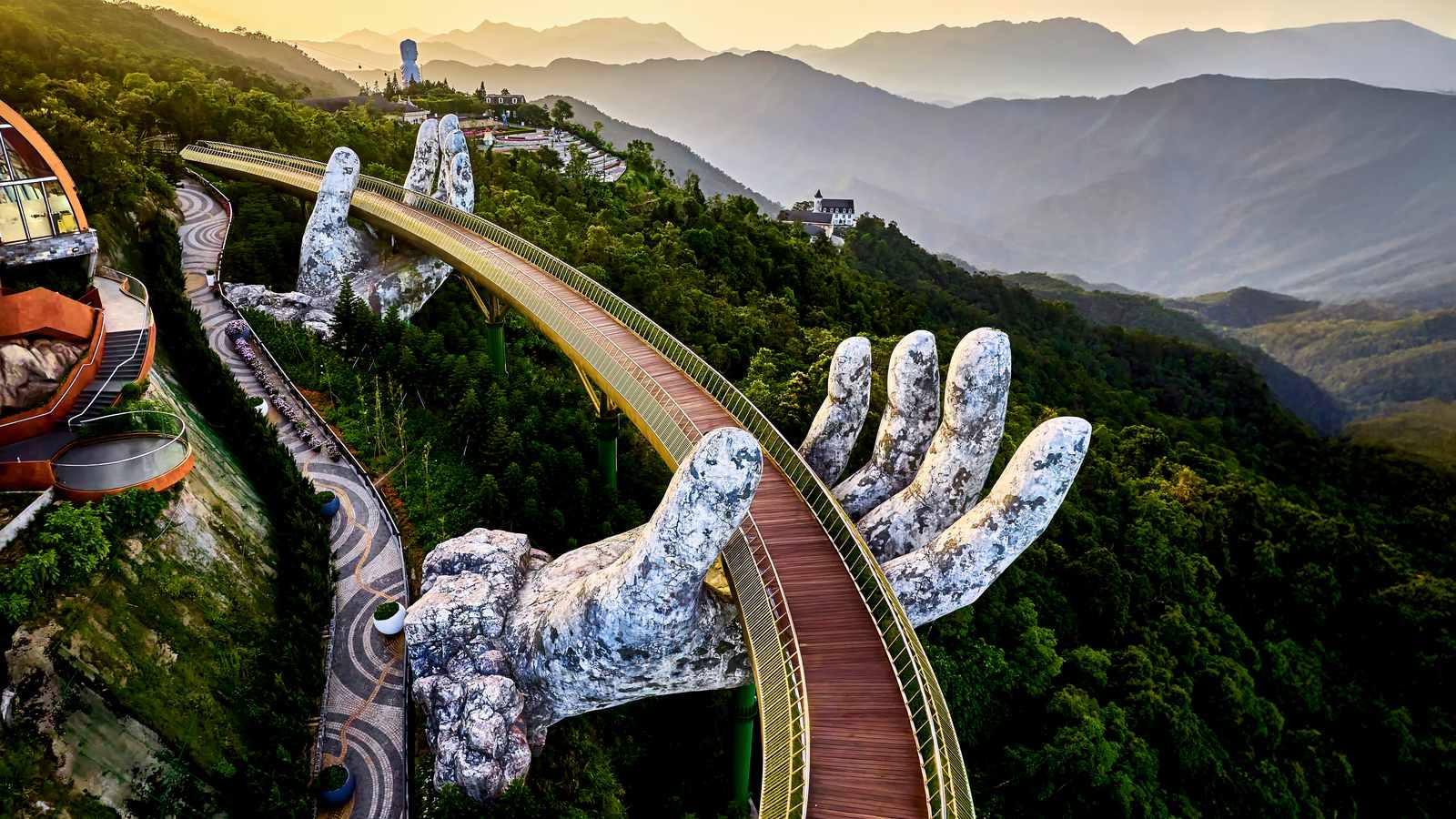 從空中俯瞰巴拿山黃金橋，兩隻巨大的石手托著金色橋身，背景是夕陽下層層疊疊的山脈
