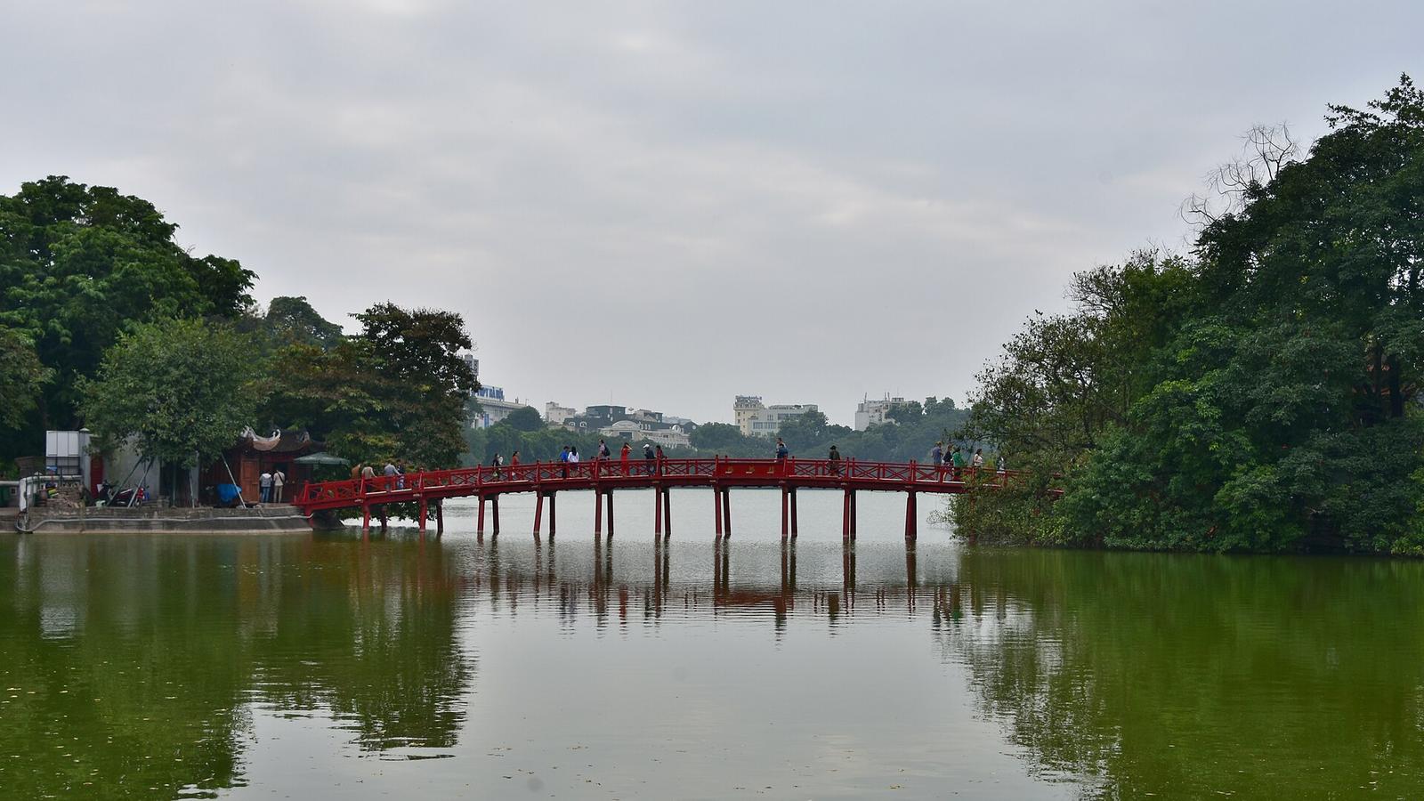 陰天下的河內還劍湖，紅色的棲旭橋橫跨湖面，兩岸濃密的綠樹倒映在水中