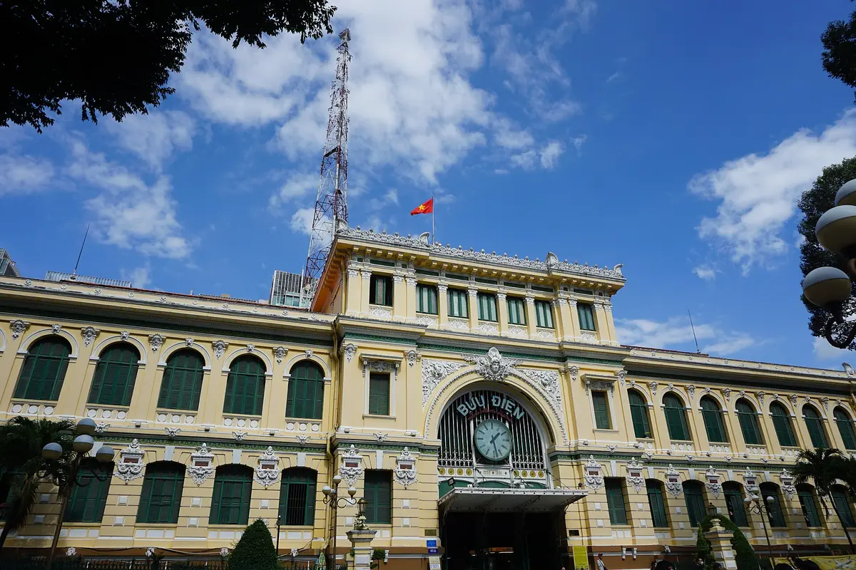 胡志明市中央郵局的黃色法式外牆，正門拱頂上有 BƯU ĐIỆN 字樣與大鐘，屋頂插著越南國旗