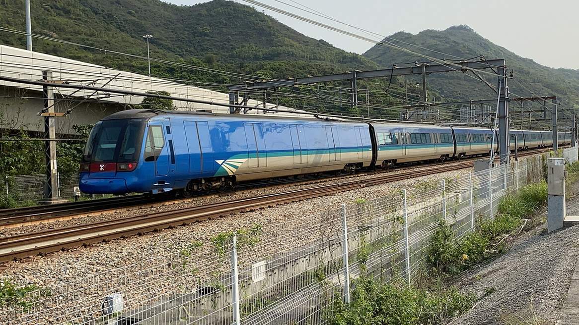 藍白塗裝的機場快綫列車行經大嶼山欣澳一帶的路軌，後方是青綠山坡