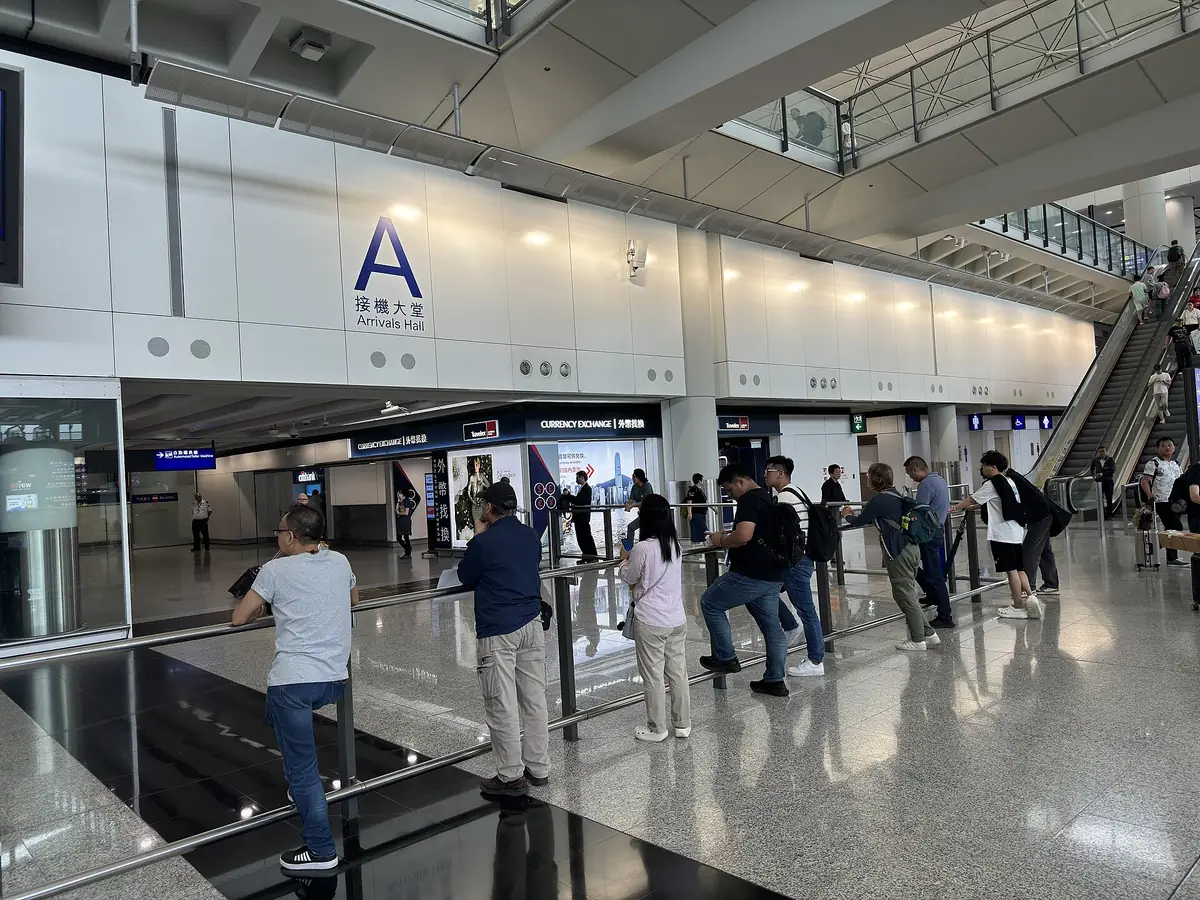 香港國際機場一號客運大樓 A 接機大堂，牆上有 A 字與接機大堂 Arrivals Hall 標示，接機民眾靠在欄杆後等候