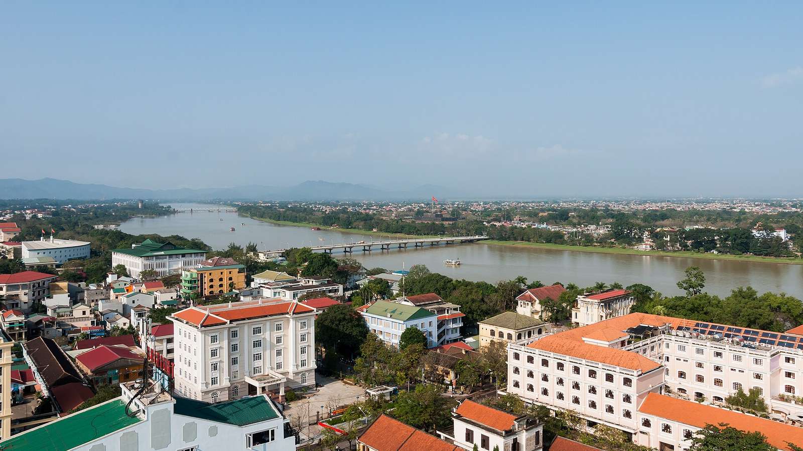 從高處看順化：香江由西往東穿過城市，河上橫著一座長橋，兩岸是低矮的街區與樹林，遠處是山
