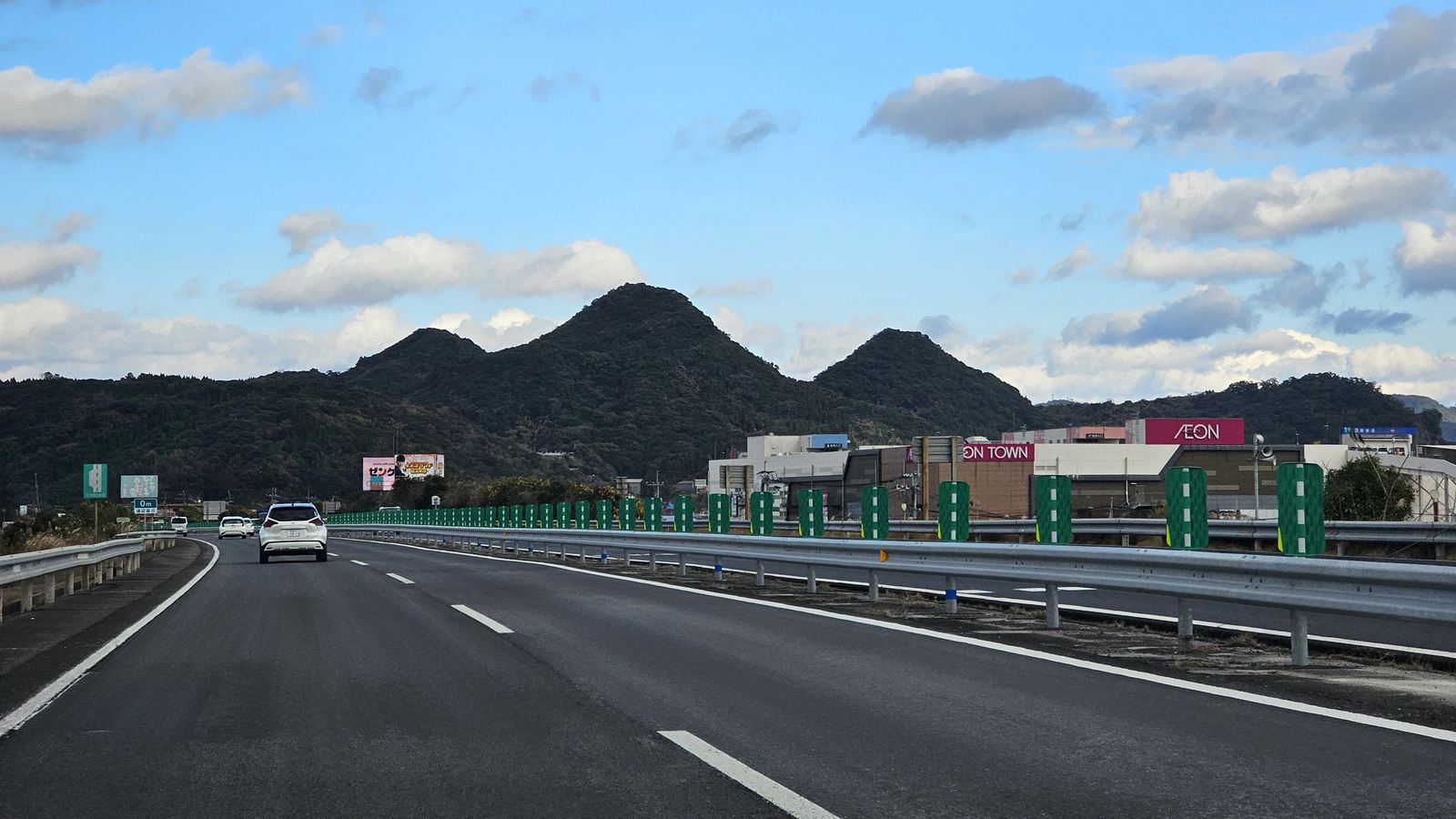 九州自動車道姶良市帖佐附近的路面，前方有幾輛轎車，遠處是山與購物中心