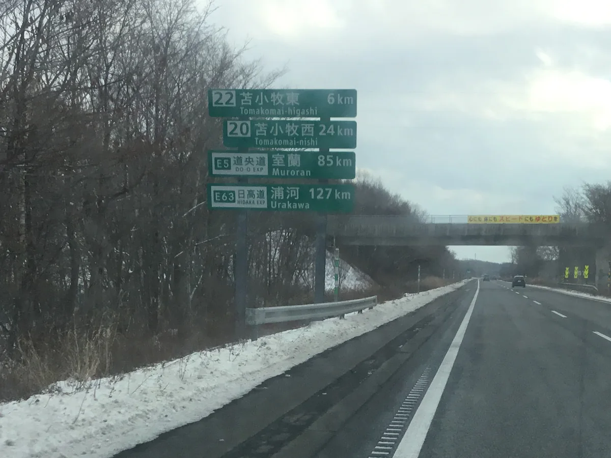 冬天的道央自動車道，路肩與樹林積雪，綠色標誌寫著苫小牧東、苫小牧西、室蘭與浦河的距離