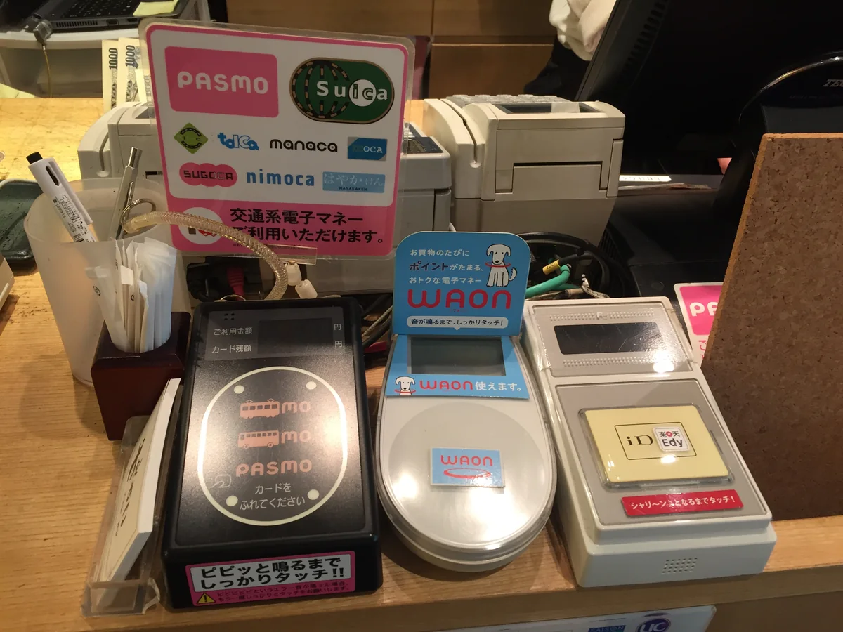 餐廳收銀台上並排的電子錢包感應機，貼著交通系 IC 卡、WAON、Edy 等標誌