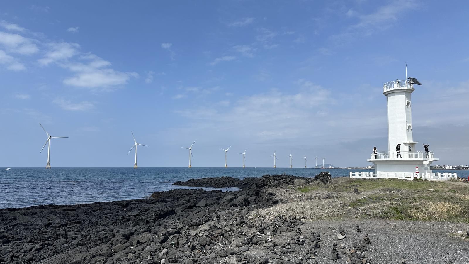 White lighthouse, basalt coast and offshore wind turbines beside Jeju's Sinchang Windmill Coastal Road