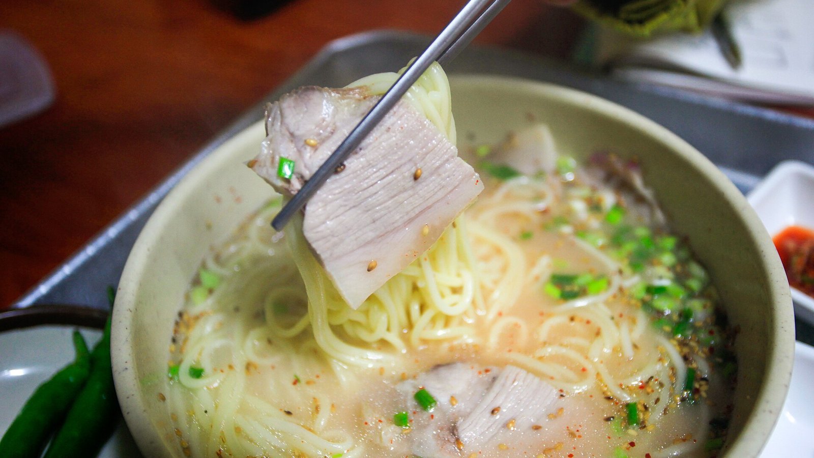 筷子夾起肉片與麵條，下方湯碗裡可見乳白湯汁、蔥花與其他肉片