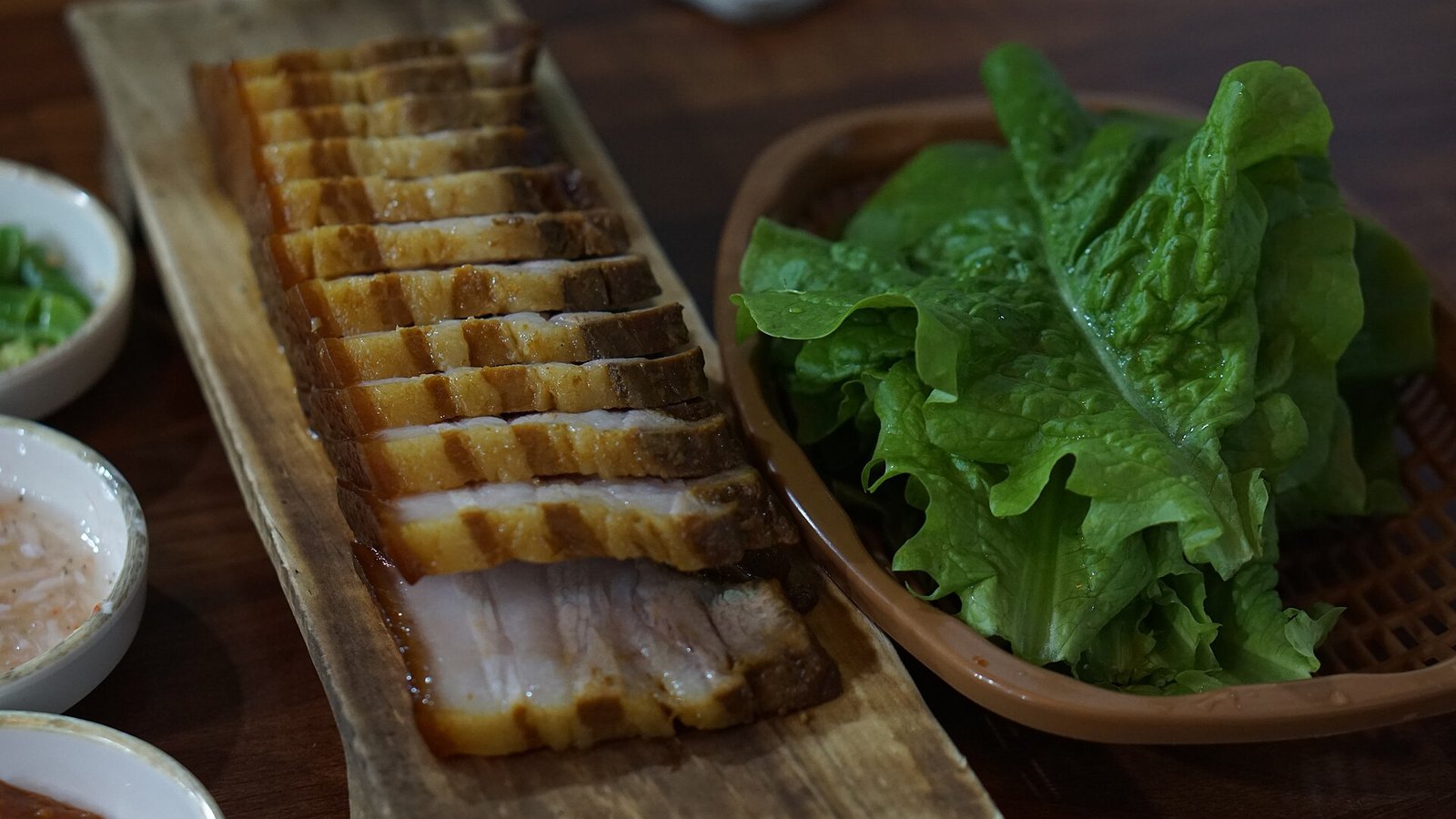 長條木砧板上排著一片片切好的豬肉，切面帶油脂與瘦肉層、外緣呈深褐色，右側是一籃新鮮生菜，左側擺著幾碟沾醬
