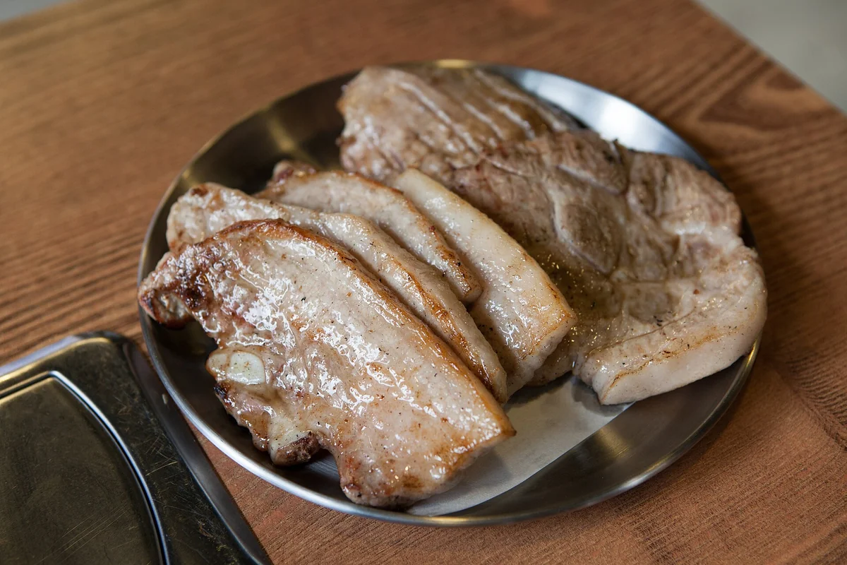 金屬盤裡盛著數片烤好的厚切五花肉，旁邊是木桌；照片無法辨認豬的品種