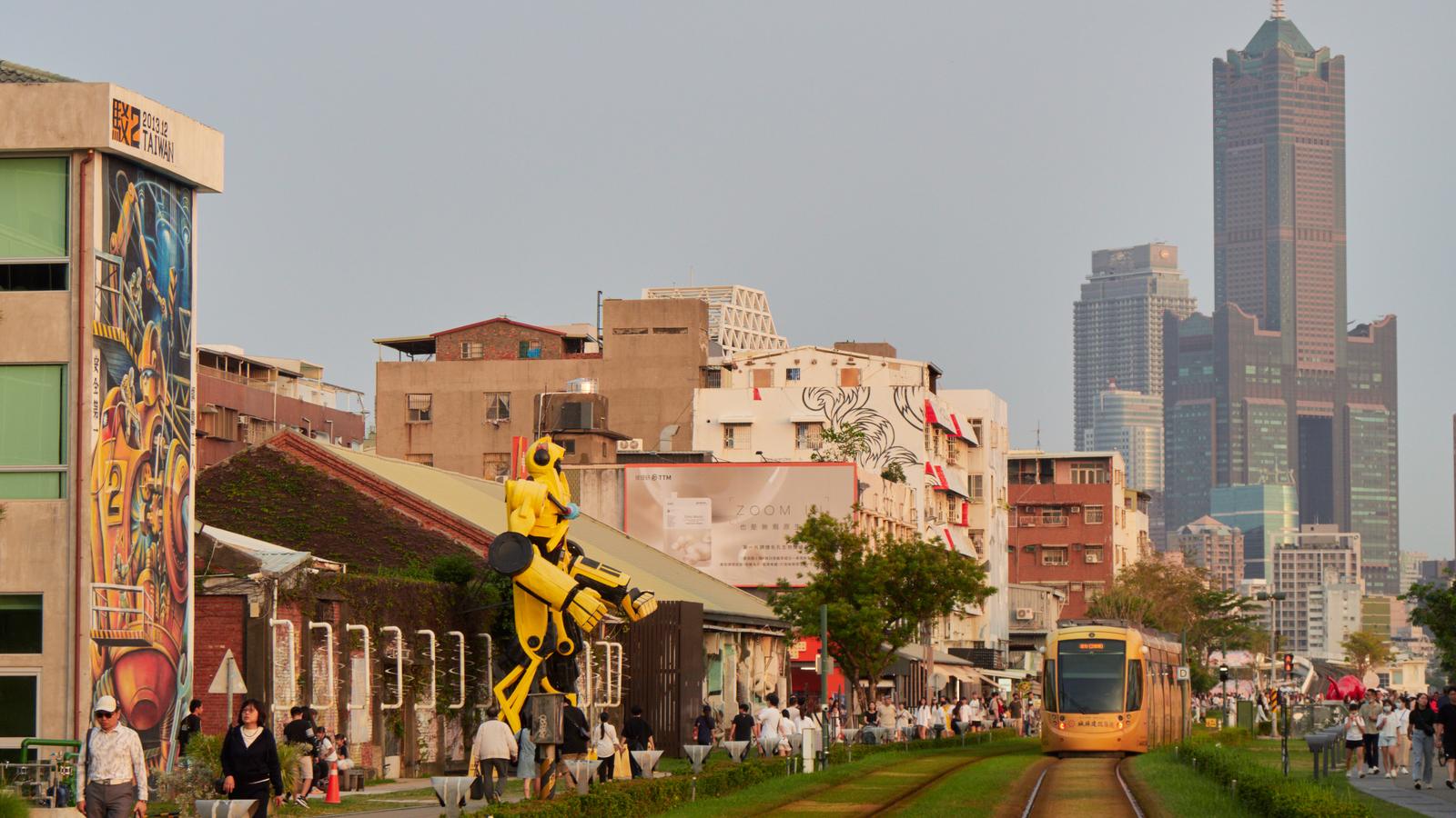 A Kaohsiung light rail tram at the Pier-2 Art Center, with a yellow robot sculpture and the 85 Sky Tower behind