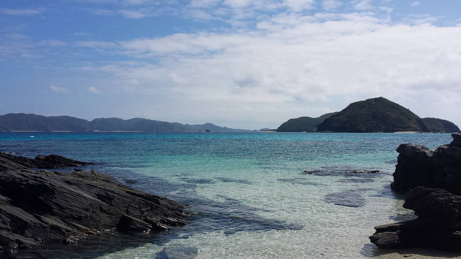 從座間味島的岩岸看出去的慶良間海域，海水由淺綠轉深藍，遠方是幾座綠色的島