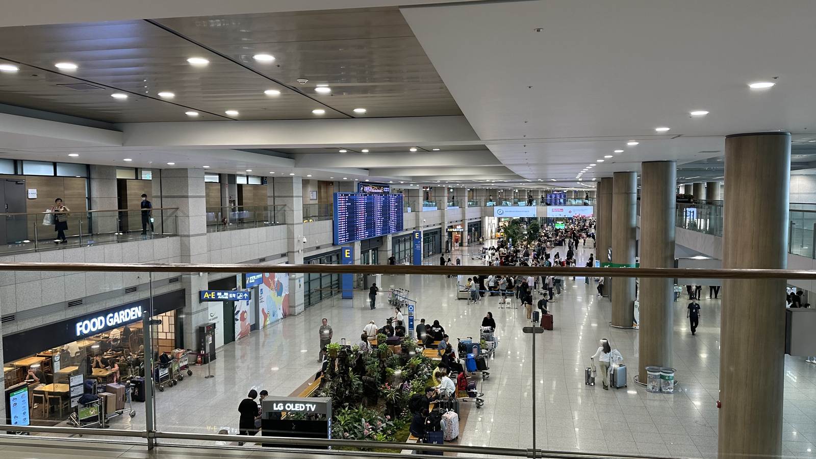 仁川機場第 1 航廈 1 樓入境大廳，旅客拖著行李走過中央的植栽區與航班看板