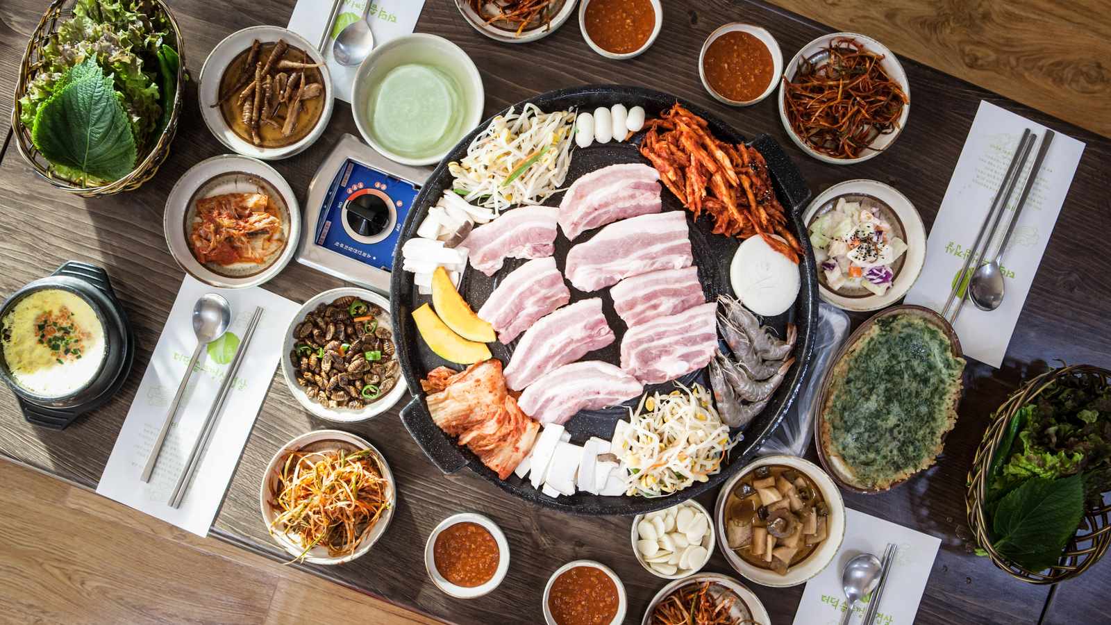 俯拍的韓國烤肉桌：中央圓形烤盤上放著五花肉、泡菜與豆芽，四周圍著十幾碟小菜、生菜籃與包飯醬