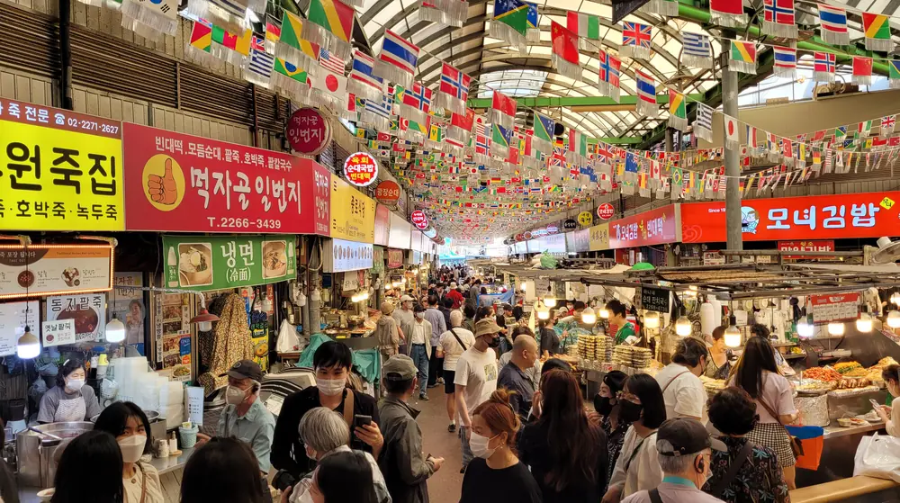 廣藏市場美食街的走道：兩側是掛著韓文招牌的小吃攤，天花板吊滿各國國旗，走道擠滿逛市場的人