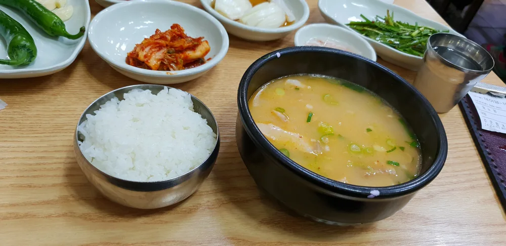 釜山的一碗豬肉湯飯：黑色石鍋裝著乳白色豬骨湯與蔥花，旁邊是一碗白飯、泡菜、青辣椒與韭菜小菜