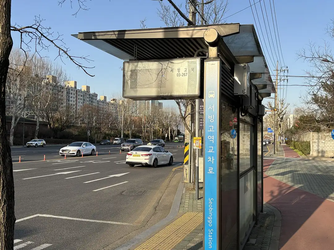 A bus shelter at an intersection by Seobinggo Station in Seoul, with the Korean stop name written vertically on a blue sign and its English name below; a white taxi passes on the road