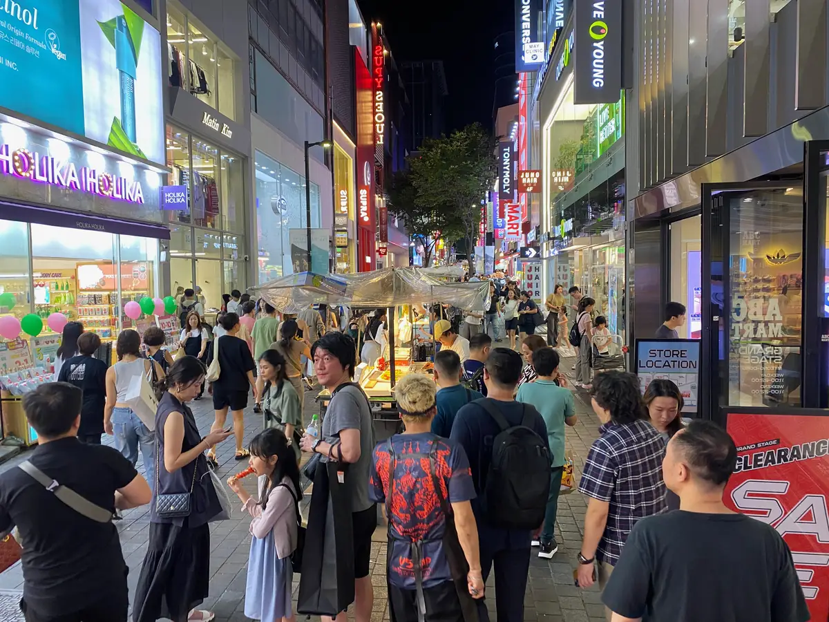 明洞夜晚的購物街，人潮穿過小吃攤，兩側是美妝店與 Olive Young 的招牌