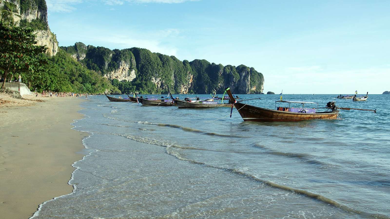 奧南海灘的沙灘與淺海，岸邊停著一排木造長尾船，左側是覆滿綠樹的石灰岩崖壁