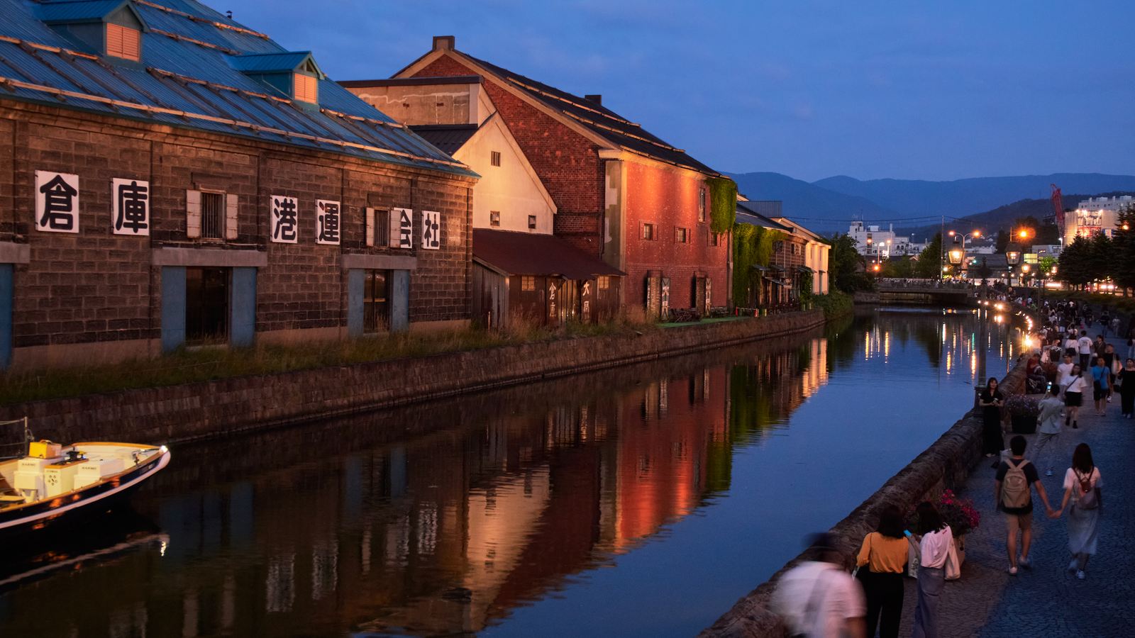 Otaru Canal lit at dusk, with stone warehouses on the left bank and visitors walking on the right