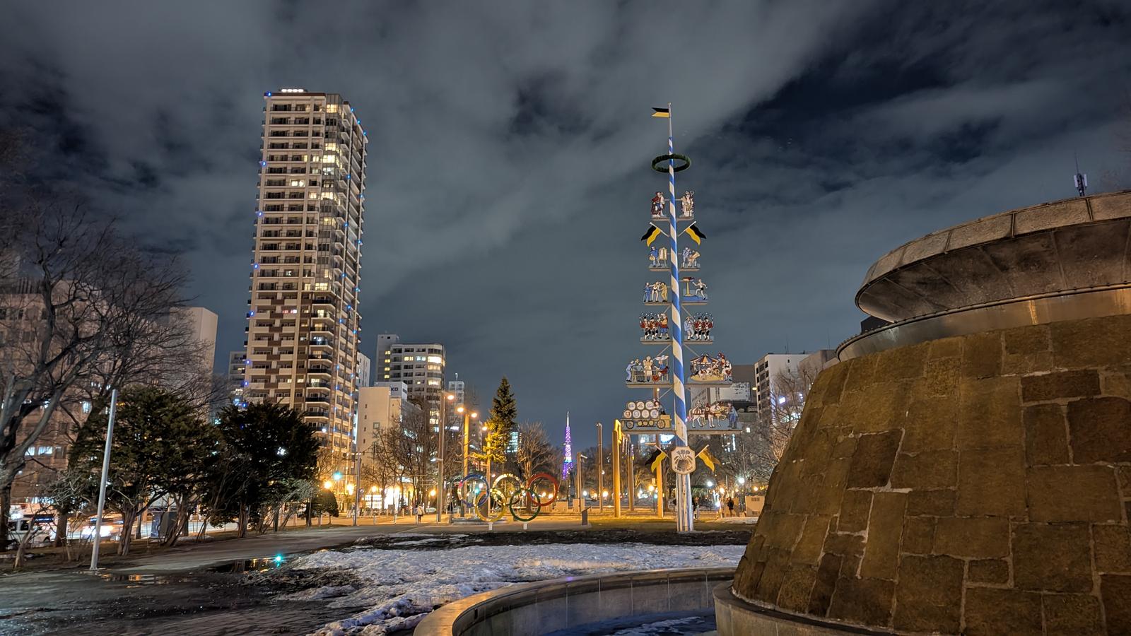 Odori Park in Sapporo at night, with illuminated buildings, a decorative pole, wet pavement and patches of snow