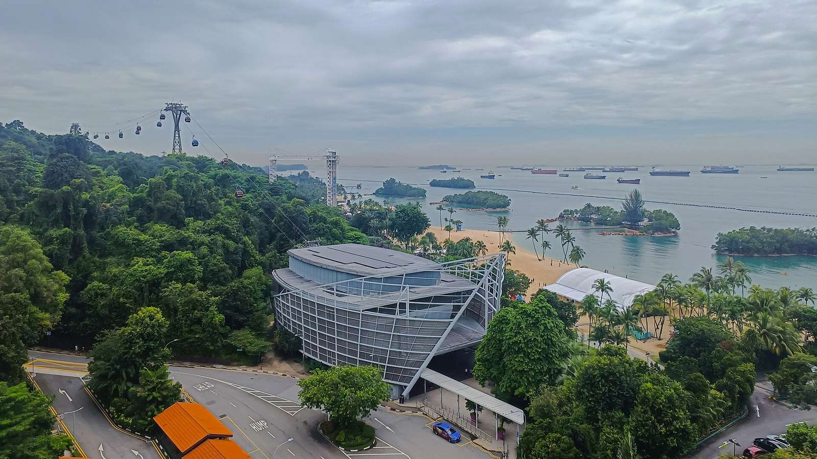 從高處俯瞰聖淘沙：左邊樹林上方是一排纜車，中間是玻璃帷幕的建築與環島道路，右邊是椰子樹與沙灘，外海停著一列貨船