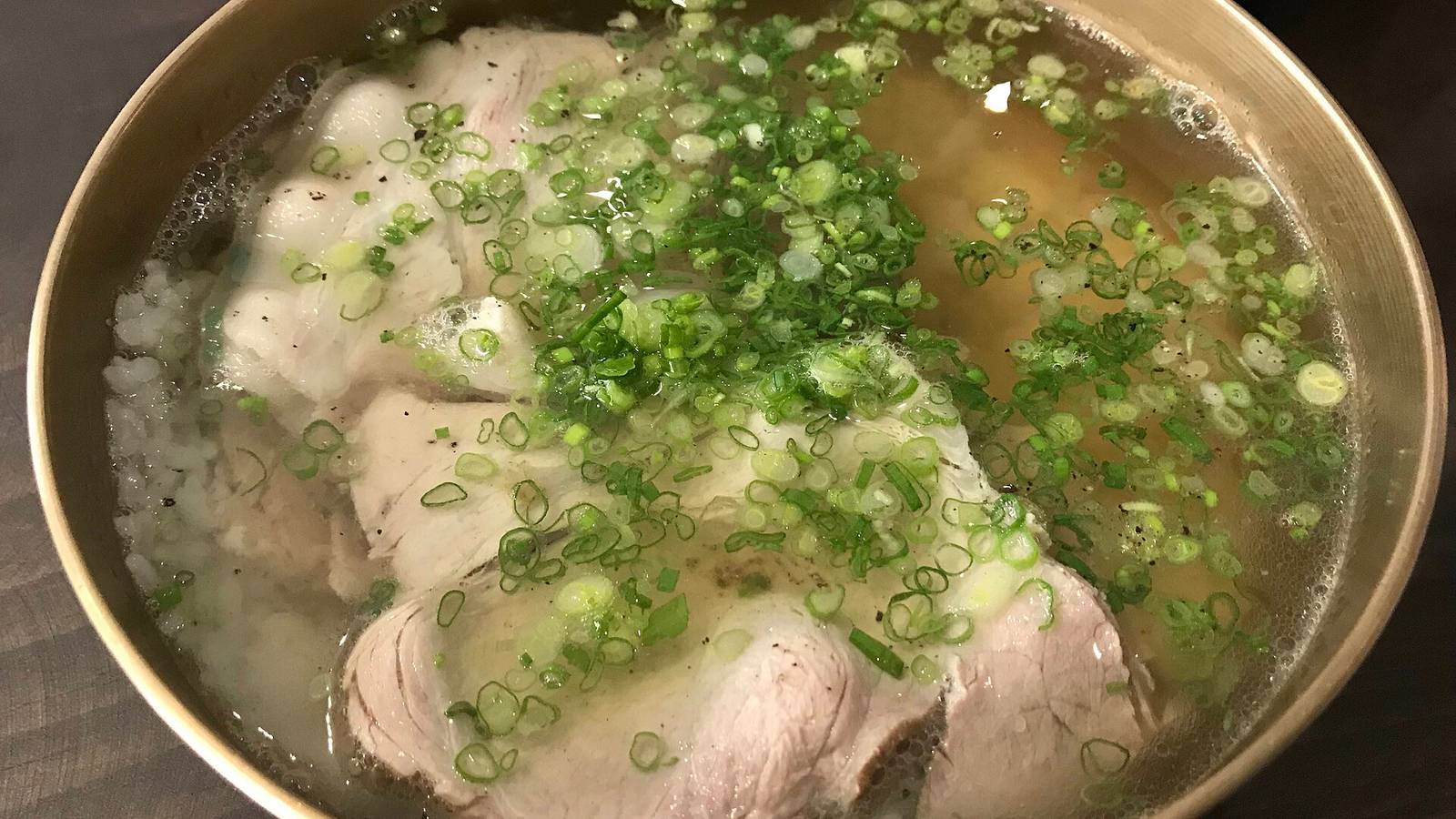 金屬碗裝著清澈的豬骨湯，湯面鋪滿蔥花，幾片白灼豬肉浮在湯裡，碗的左側露出一點白飯
