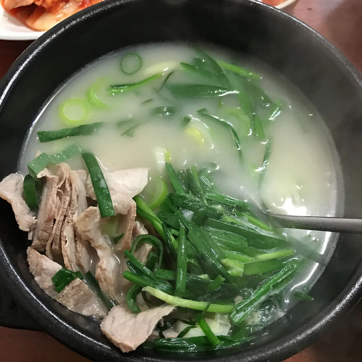 黑色陶碗裝著乳白色豬骨湯，湯面鋪滿切段的韭菜與蔥花，左下角看得到幾片豬肉，一支湯匙從碗的右側伸入湯中