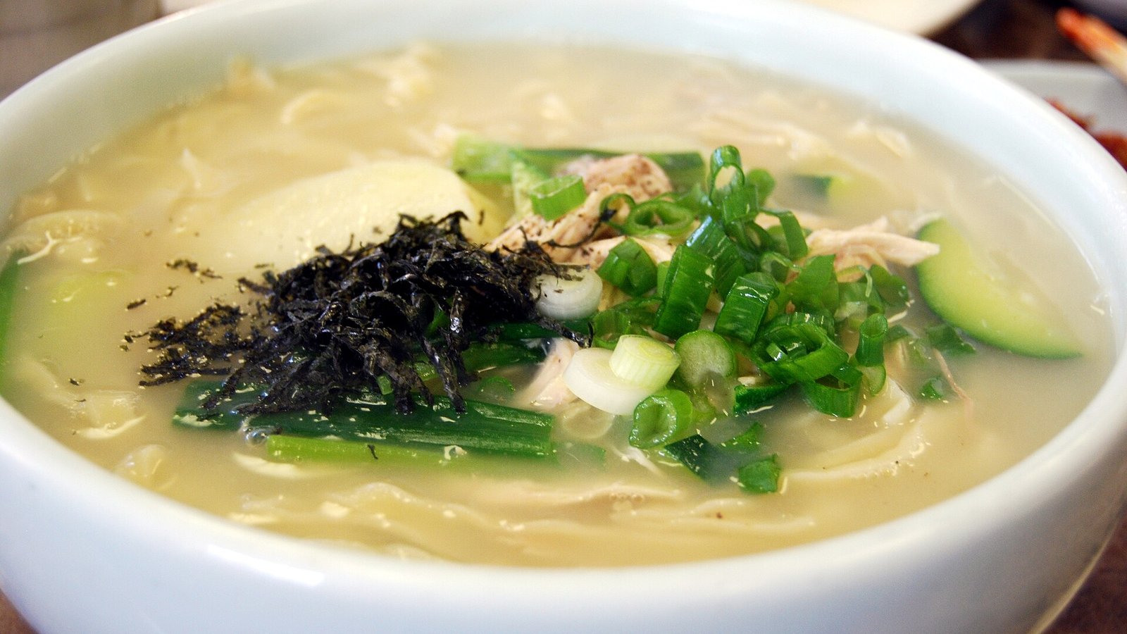 白色圓碗裡的湯麵,湯色淡黃微濁,鋪著切碎的黑色海苔絲、蔥花與撕成細絲的淡色肉,還有綠皮的瓜片,後方模糊處是幾碟其他小菜