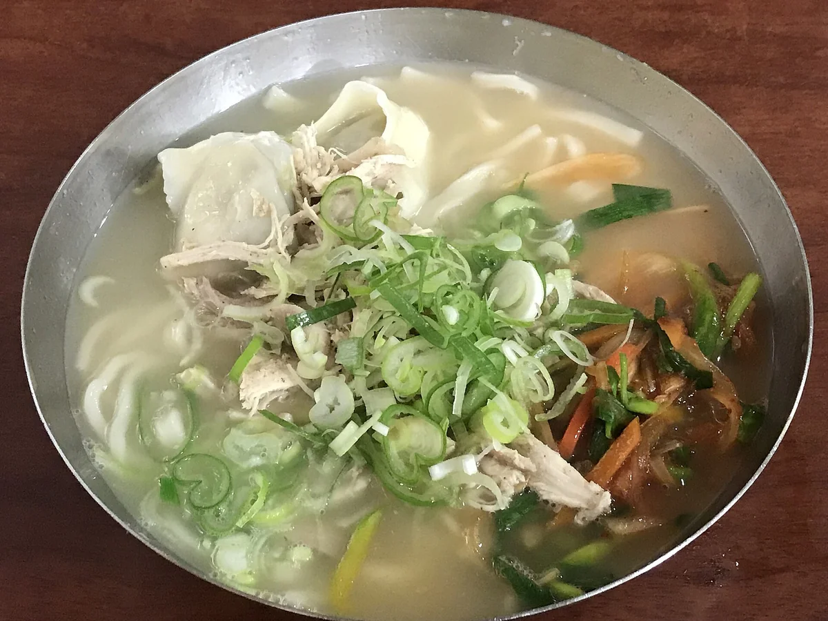銀色不鏽鋼淺碗裝著湯麵，左側是一顆摺邊明顯的大水餃與撕成絲的雞肉，中間堆著捲曲的蔥絲，右側另外盛著一小份橘紅色的拌菜