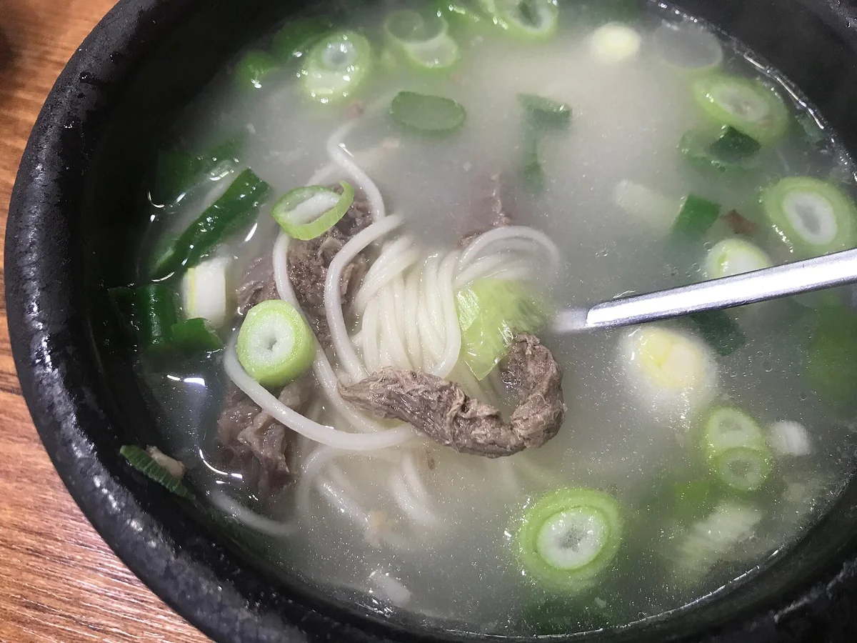 俯拍一碗雪濃湯，湯裡有細麵、數片牛肉與蔥花，一根細長的金屬餐具從右側伸入碗中