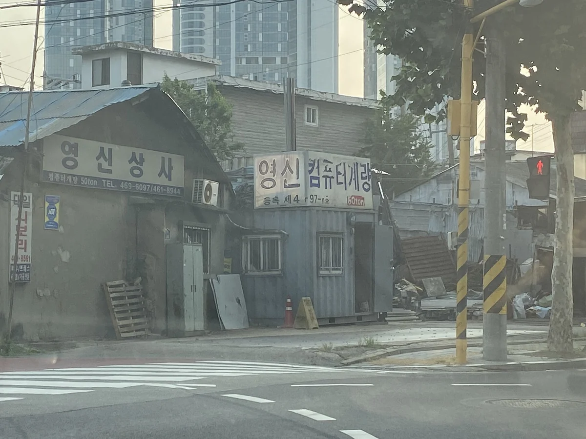 聖水洞街角的低矮舊建物，鐵皮屋頂下掛著幾面韓文招牌，後方是高聳的現代高樓，前景是行人穿越道