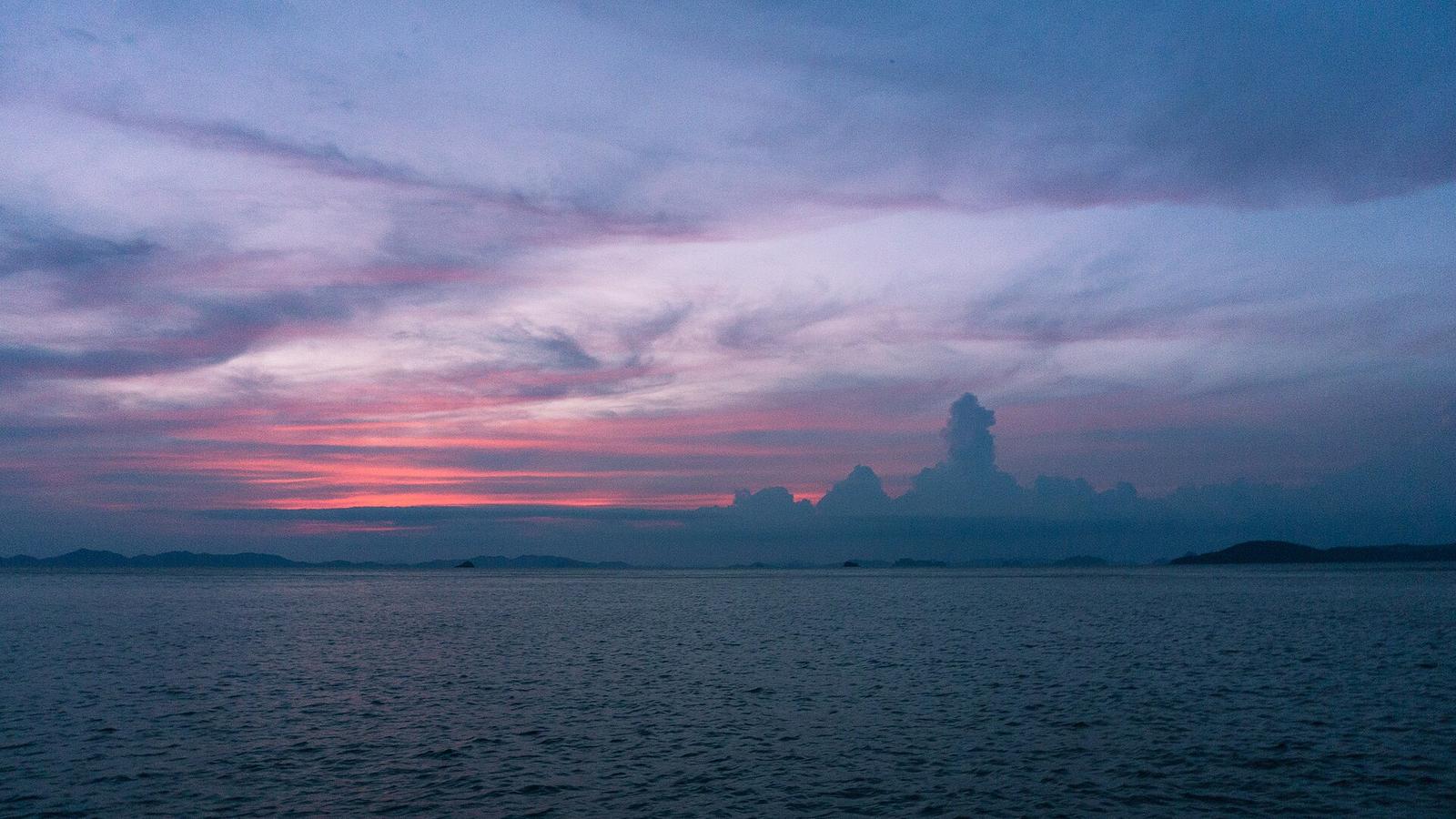 安達曼海的傍晚海面，天空是一整片灰紫色雲層、靠近地平線透出橘紅色霞光，遠方有低矮的島影與一朵高聳的積雲