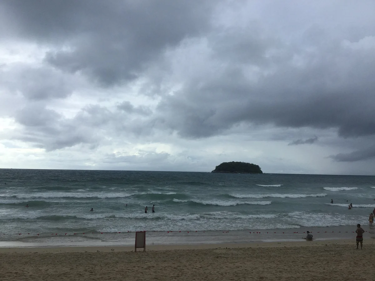 普吉卡塔海灘的陰天景象，天空滿是厚重灰雲，海面起浪、白色浪花一排排打上沙灘，岸邊拉著一條紅色旗繩