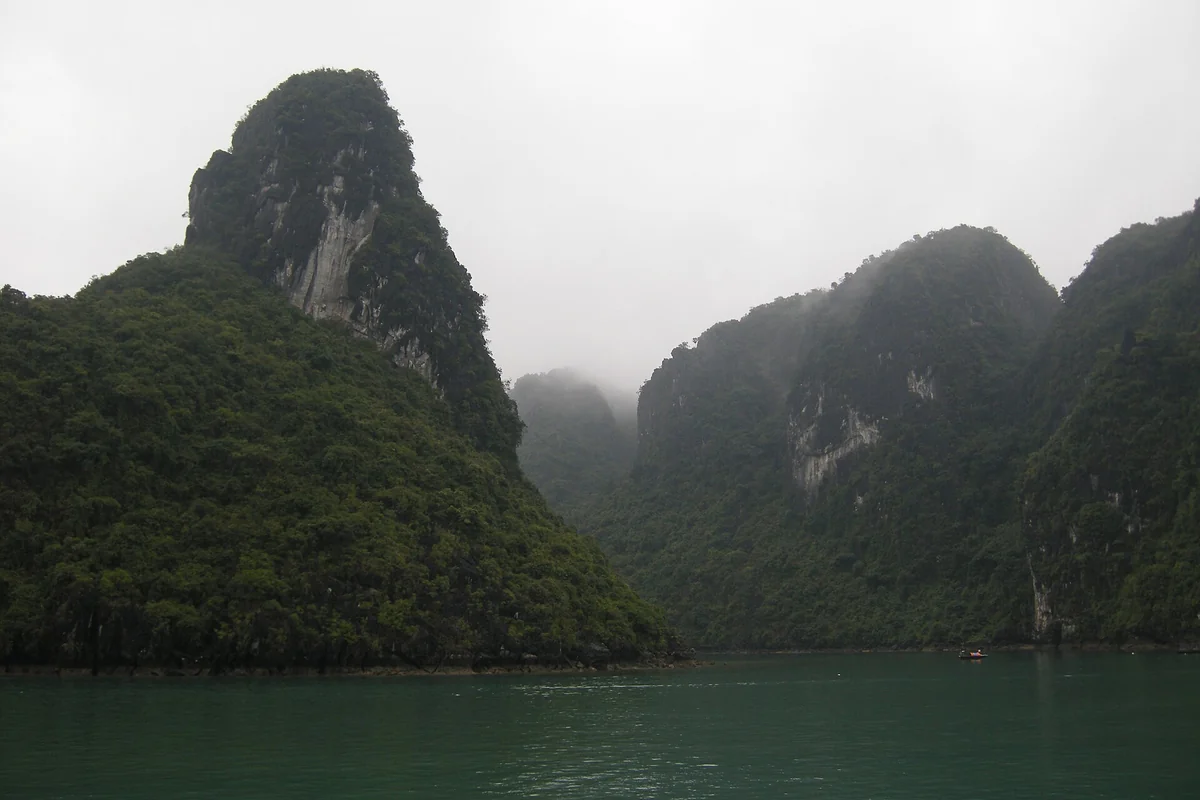 下龍灣的石灰岩島嶼籠罩在灰白色濃霧裡，海面平靜，遠處有一艘小船