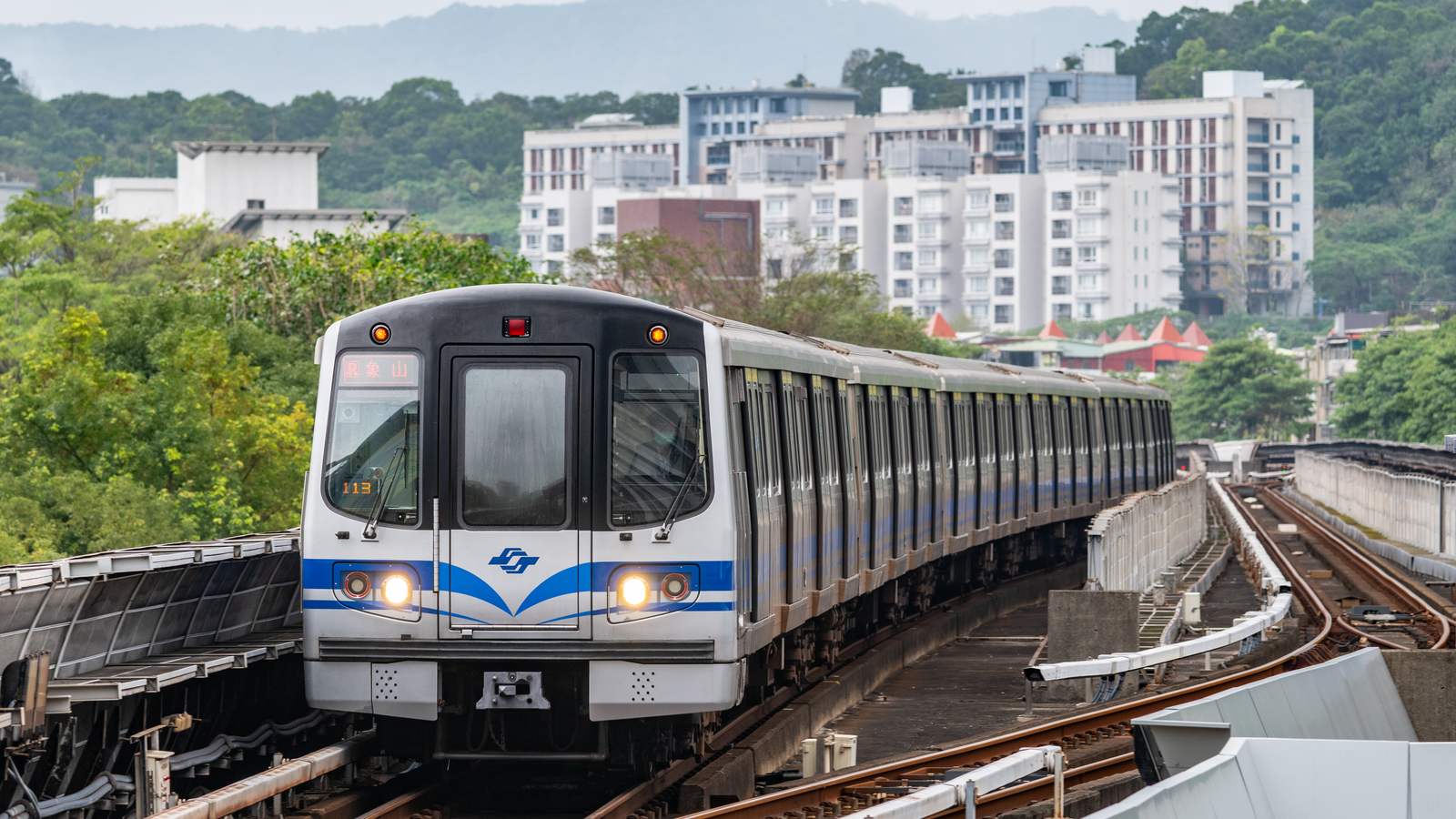 A Taipei Metro Tamsui-Xinyi Line train with blue stripes on the elevated track at Shipai station, green hills behind