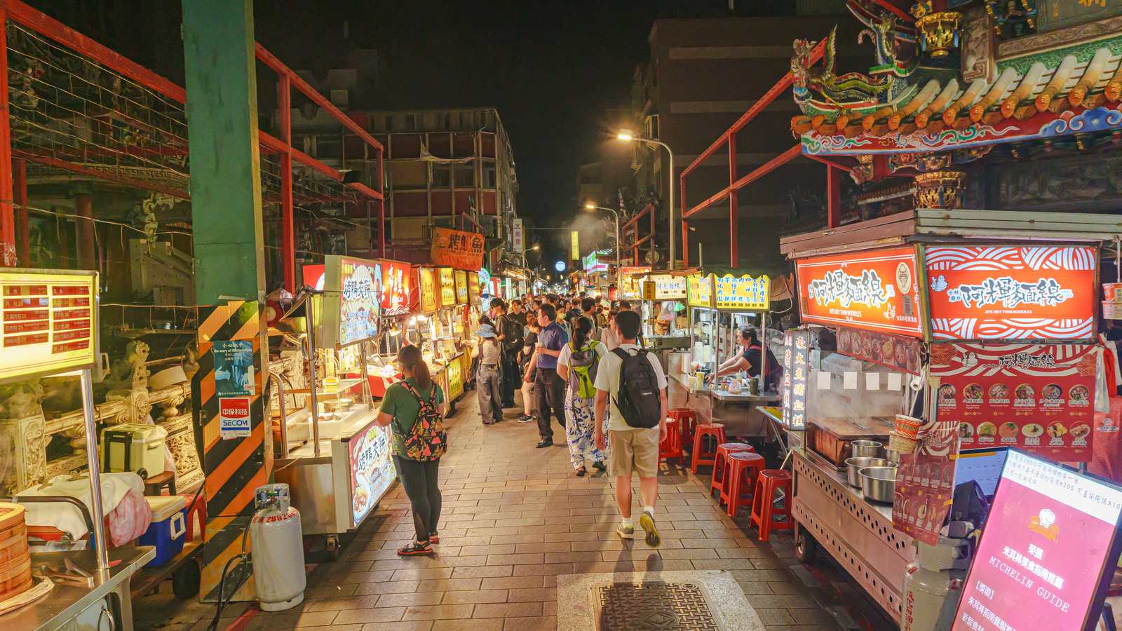 Night view of Danan Road in Shilin Night Market: a paved lane of lit food stalls and red stools beside a temple roof
