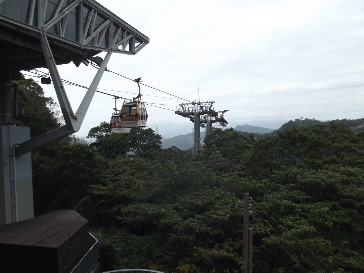 A Maokong Gondola cabin leaving Maokong Station above wooded hills, with Taipei 101 small on the horizon