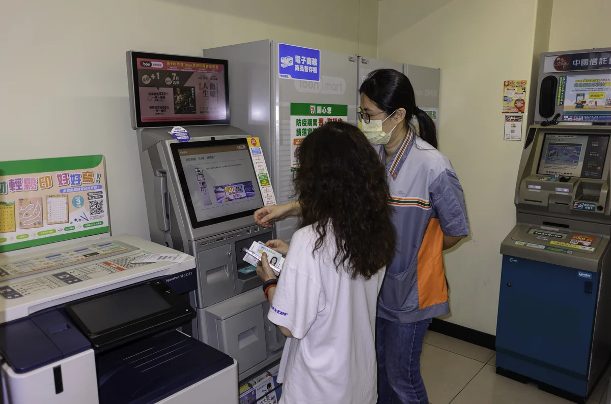 A 7-ELEVEN staff member helps a customer at the ibon kiosk inside a Taipei store, with a bank ATM behind them