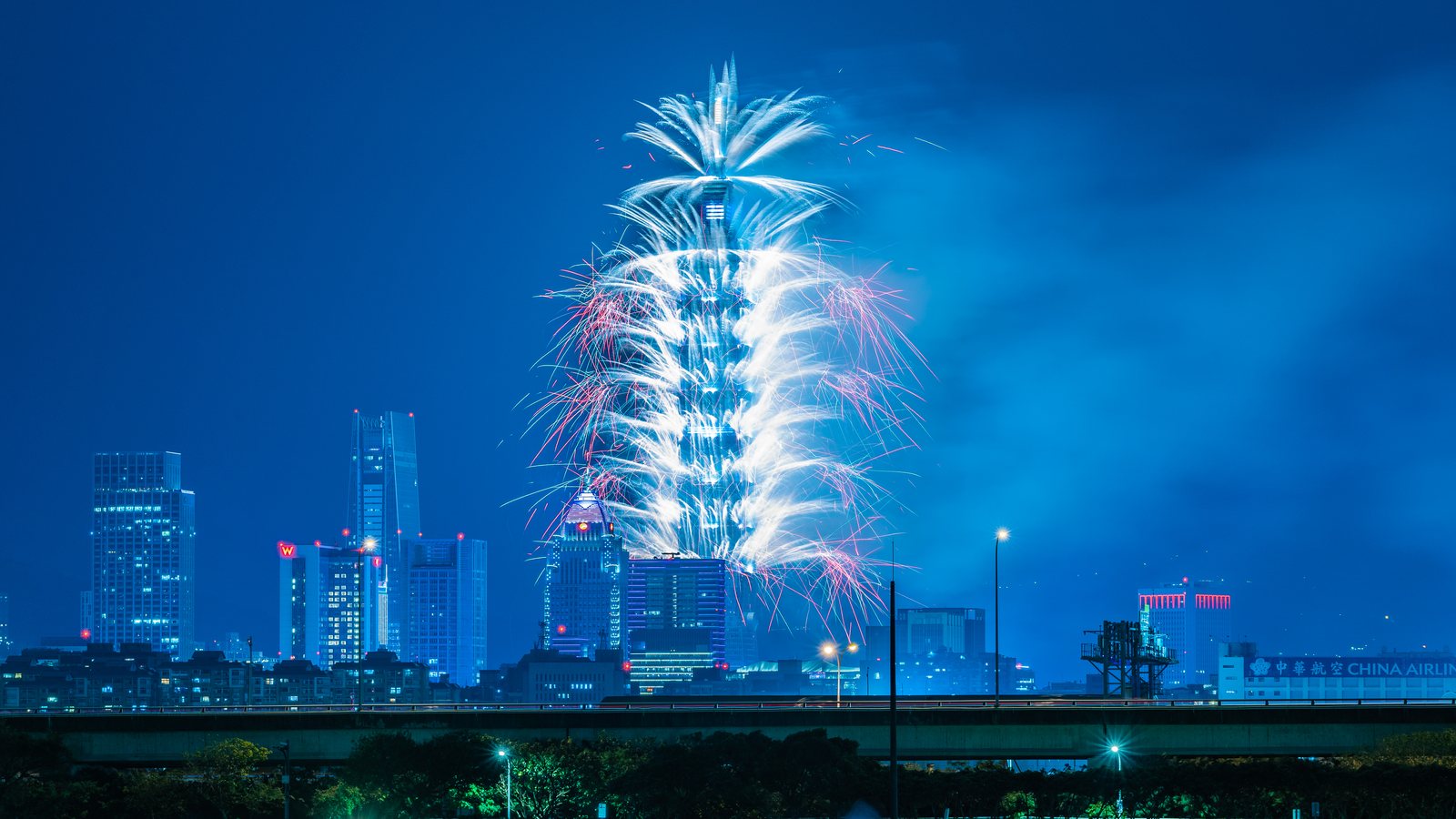 Fireworks pour down the sides of Taipei 101 above the night skyline, seen from a riverside park