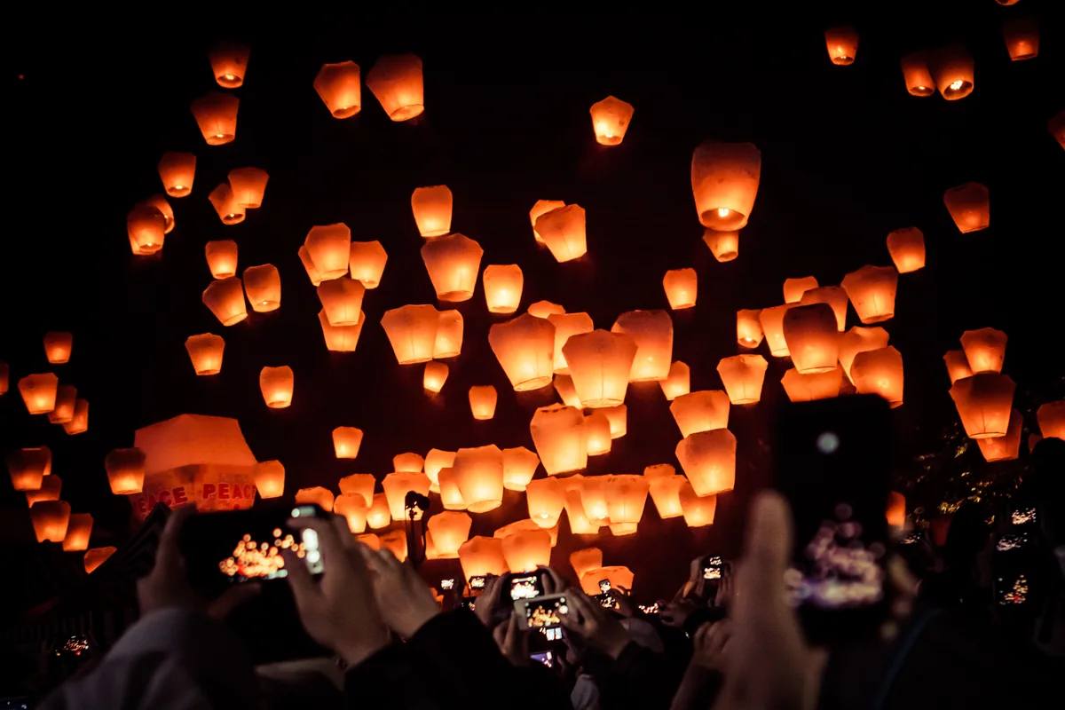 Hundreds of glowing sky lanterns rising into the night sky over a crowd holding up phones at Pingxi