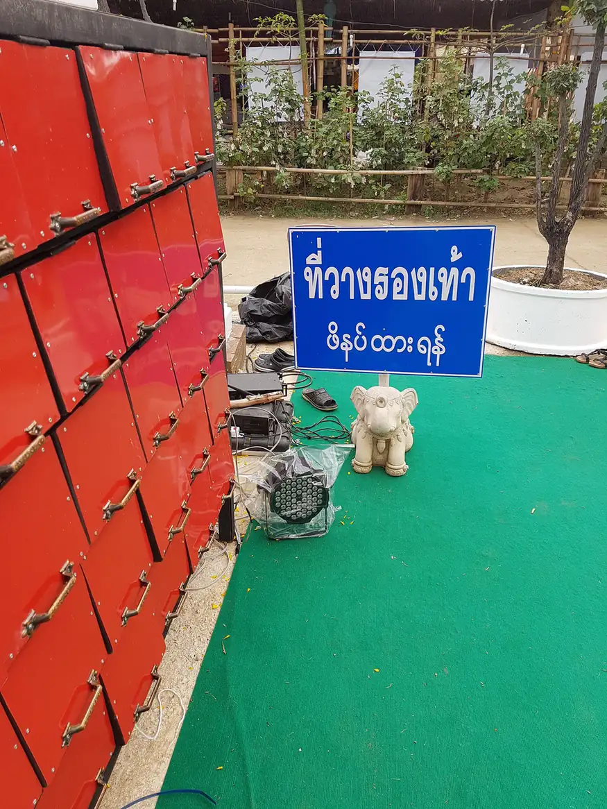 寺廟入口旁成排的紅色鞋櫃，旁邊立著藍底白字的放鞋處告示牌與一尊小象雕像，地上鋪著綠色地毯