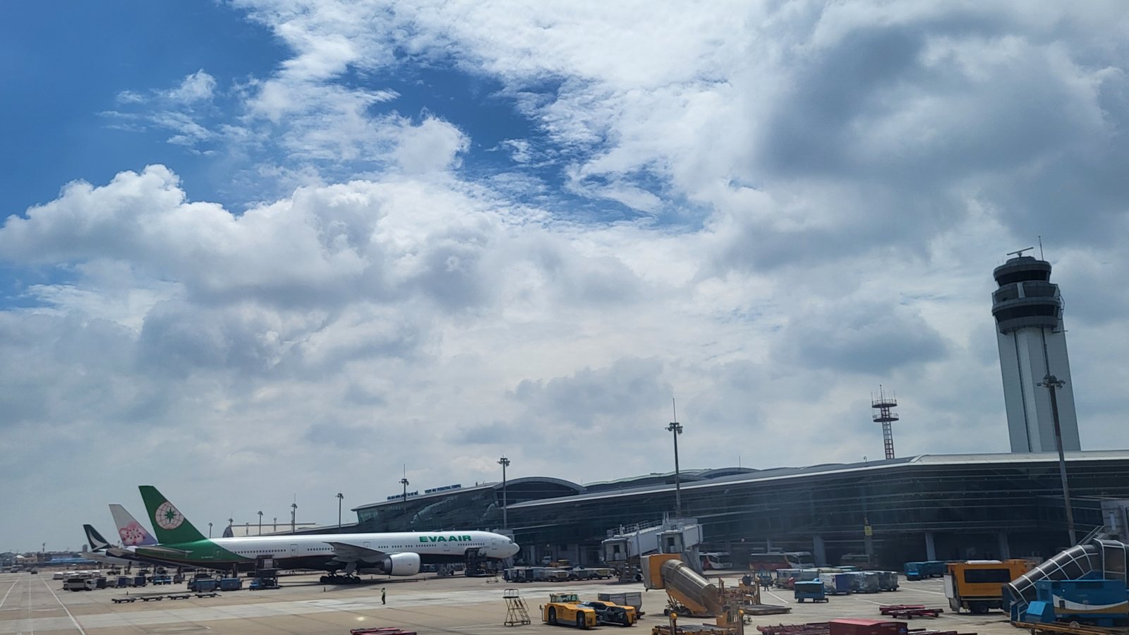 胡志明市新山一機場國際線 T2 航廈前的停機坪，一架長榮航空客機停在航廈旁，後方有其他客機尾翼，右側是航廈與塔台，天空多雲