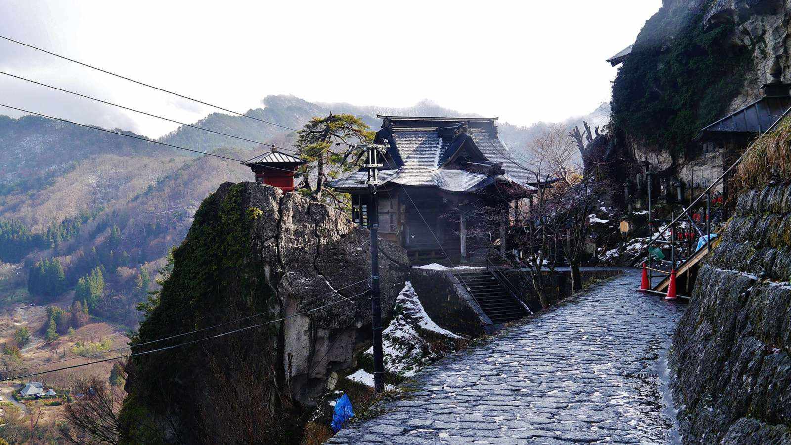 山寺立石寺的開山堂與岩頂上的紅色納經堂立在崖邊，石板參拜道沿著山壁往上，遠方是山谷與層疊的山稜